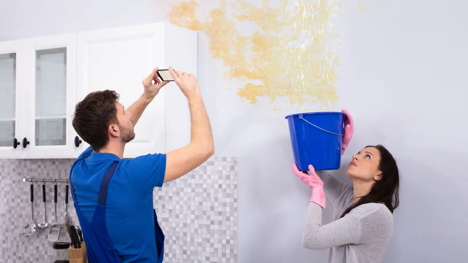 Homeowners photographing a leaking ceiling stain while catching drips in a bucket